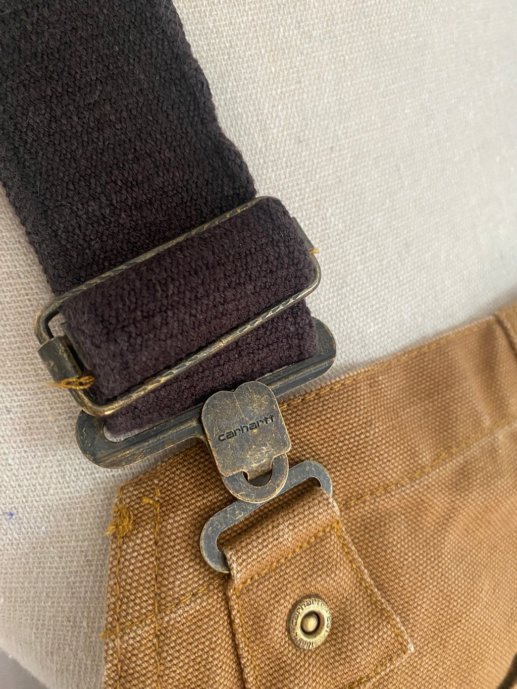 Détail fermeture de bretelle Carhartt, boucle métallique bronze gravée et système d'attache réglable